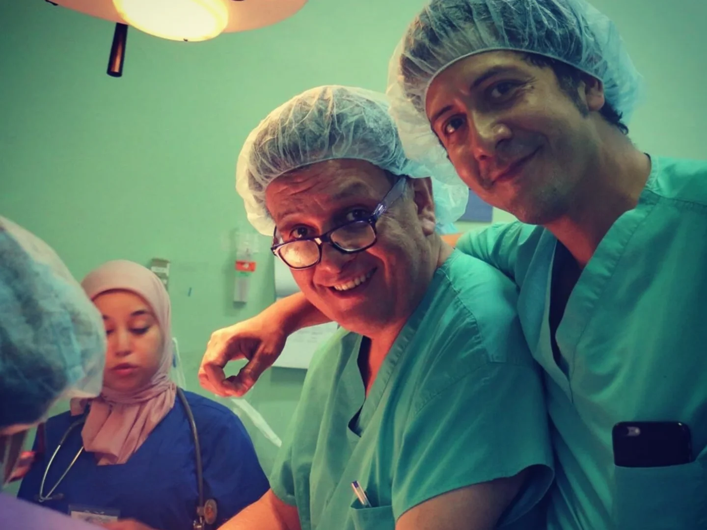 Dr. Nicolas Sierra takes a selfie-style photo with his father, Dr. Plinio Sierra. They are smiling at the camera while wearing green scrubs and disposable hair covers in an operating room.